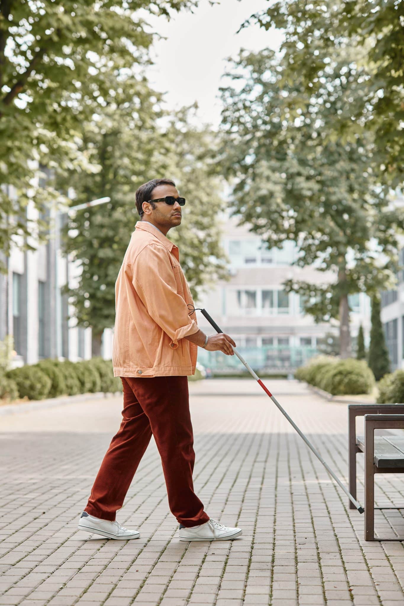 blind man with glasses and walking stick in orange jacket walking in park